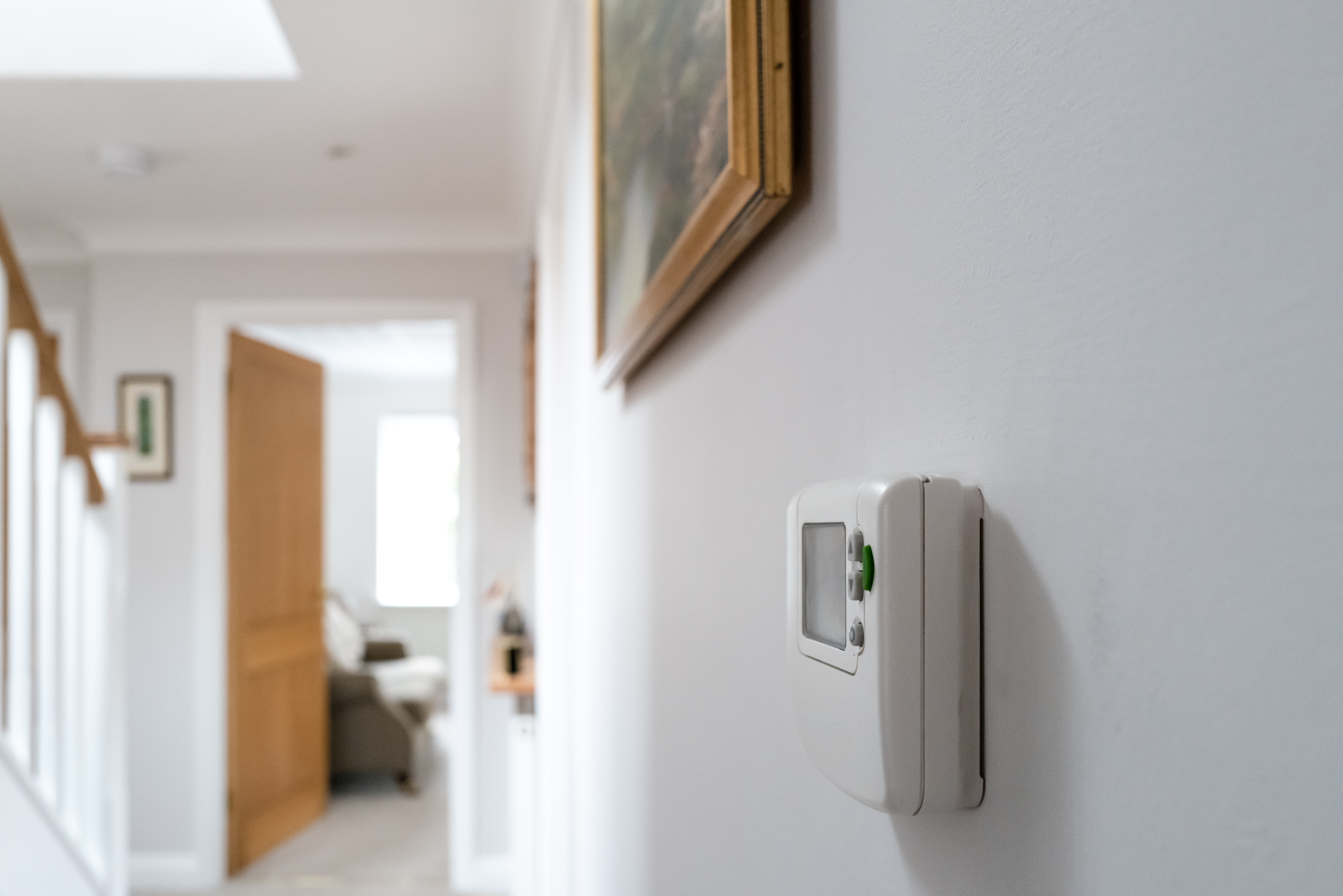 White hallway with a digital thermostat on the wall. A wooden staircase and an open door leading to a bright room create a calm and inviting atmosphere.