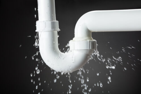 A leaking white PVC pipe with water dripping from the joints against a dark gray background. Water droplets are visibly splashing around the pipe, indicating a need for repair.